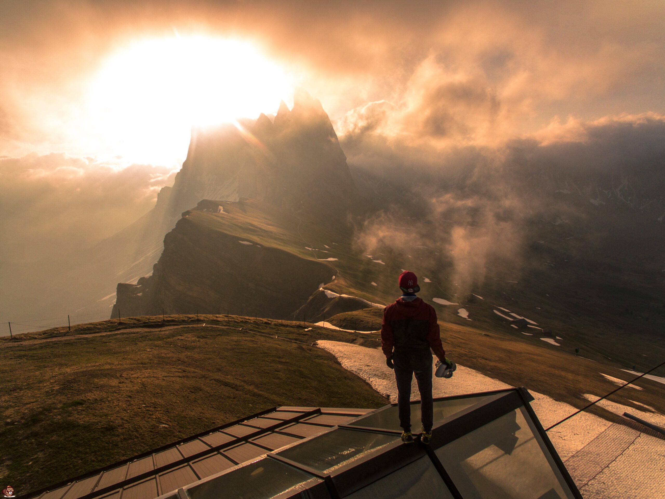 Person in red jacket and cap stands on rooftop overlooking sunlit mountain landscape with mist.