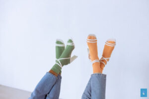 Two pairs of legs in blue jeans, green and orange socks with sandals, white strap design.
