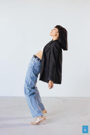 Person leaning backward in minimalist space, in black blazer, white top, jeans, and clear heels.
