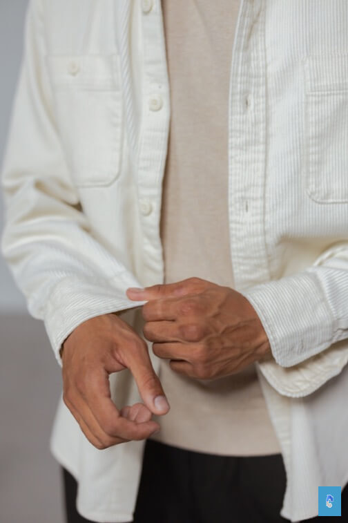 Person adjusting the cuff of a white button-up shirt over a beige top indoors.