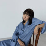 Person in blue garment seated on wooden chair, looking contemplatively to the side.