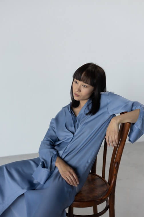 Person in blue garment seated on wooden chair, looking contemplatively to the side.
