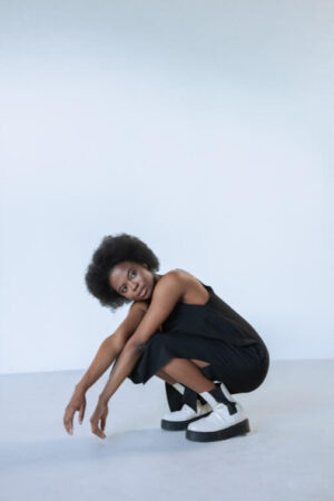 Person crouching on floor, short curly hair, black sleeveless outfit, white platform shoes.