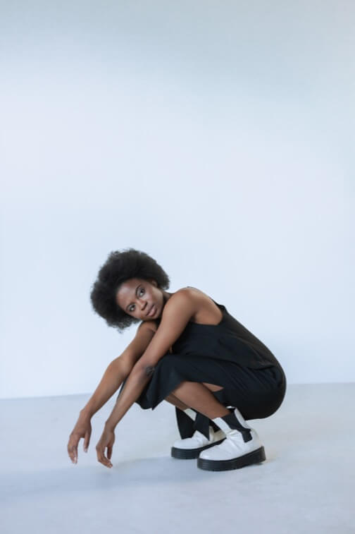 Person crouching on floor, short curly hair, black sleeveless outfit, white platform shoes.