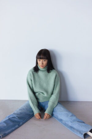 Person in light green sweater, blue jeans, sitting on floor against light wall with legs extended.