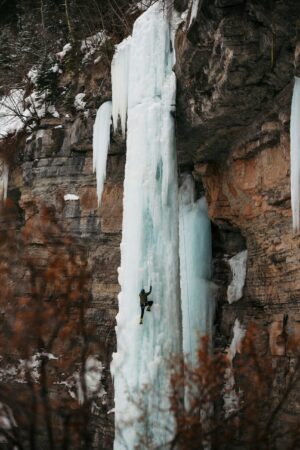 Person ice climbing a tall frozen waterfall on rocky cliffs with bare trees in winter.