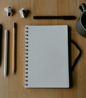 Spiral dotted notebook with pencils, pen, earbuds, and mug on wooden surface.