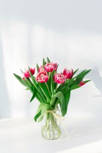 Glass jar with pink tulips and green leaves, tied with ribbon, light background.