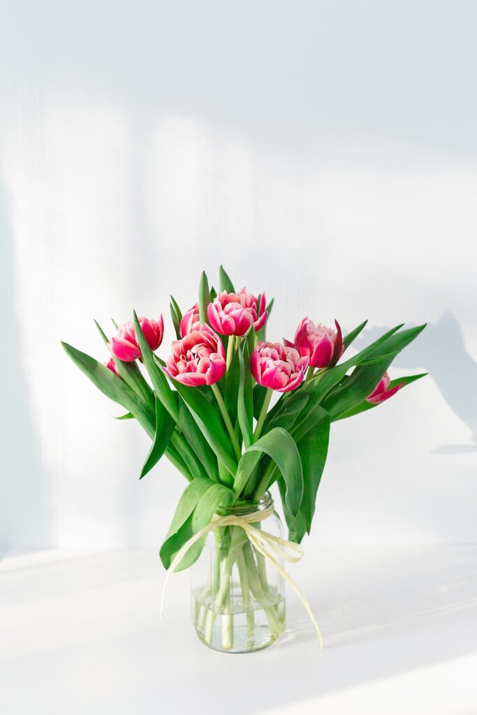 Glass jar with pink tulips and green leaves, tied with ribbon, light background.