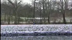 Person walking on a path beside a rocky embankment along a body of water with leafless trees and snow.