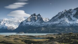 Snow-covered mountain peaks with a lake and rocky grassy foreground under a partly cloudy sky