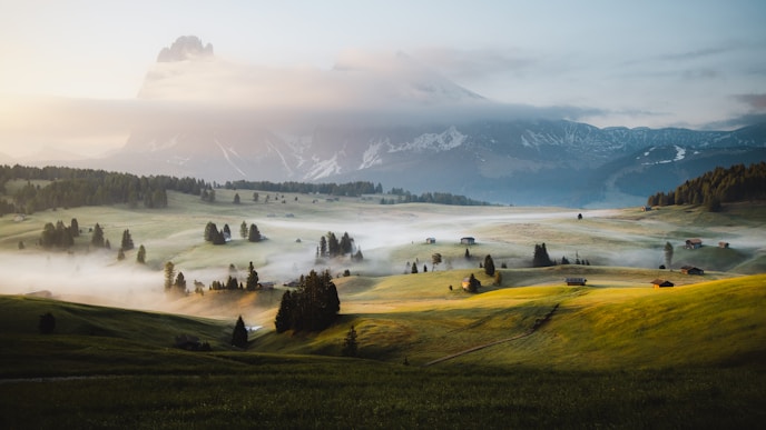 Rolling green hills with scattered trees and buildings, morning mist, and a mountain in clouds.