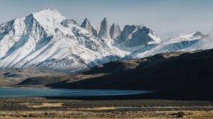 Snow-covered mountain range with rocky peaks, lake, and dry grassland in foreground.