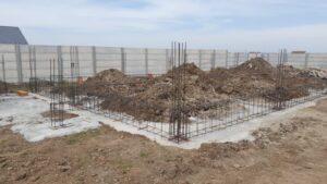 Construction site with steel reinforcement for foundation, piles of dirt, concrete base, and a concrete fence.