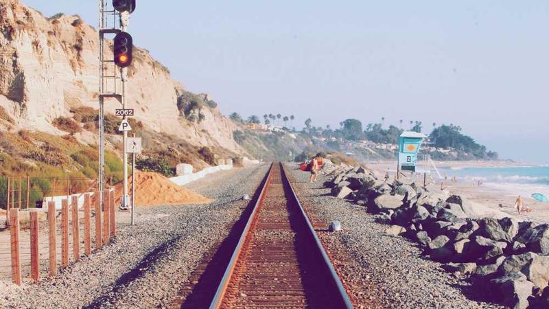 Cale ferată de coastă cu semnal roșu, stânci în stânga și plajă cu oameni în dreapta.