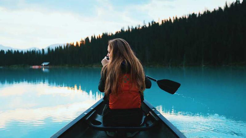 Person wearing red life jacket paddling a canoe on a calm lake surrounded by forest and hills