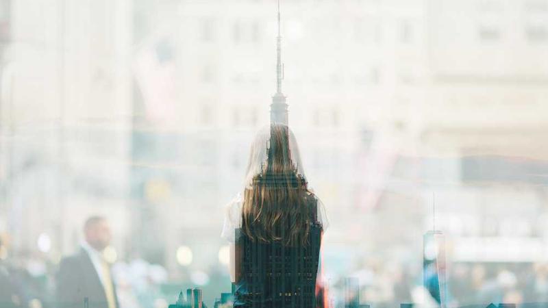 Double exposure of a woman with long hair seen from behind and a city skyline with tall buildings.